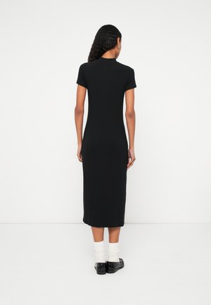 Mujer con cabello rizado lleva un vestido midi negro de manga corta, calcetines blancos y mocasines negros, de pie frente a una pared blanca.
