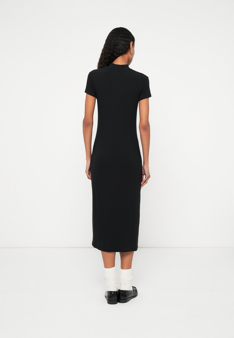 Femme aux cheveux bouclés porte une robe midi noire à manches courtes, des chaussettes blanches et des mocassins noirs, debout face à un mur blanc.