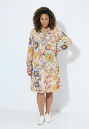 Kobieta z kręconymi włosami, ubrana w sukienkę do kolan z długim rękawem, w kolorowe kwiatowe i motylkowe wzory, stojąca z jedną ręką na biodrze.