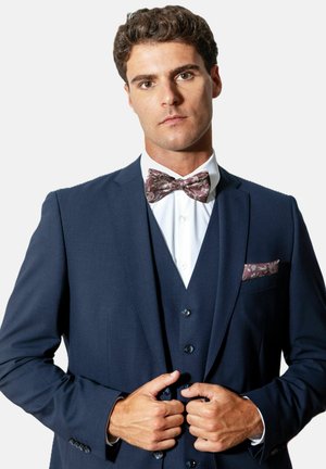 Homme ajuste un costume trois pièces bleu marine avec une chemise blanche, un nœud papillon marron à motifs et un mouchoir assorti sur fond uni.