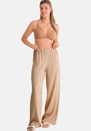 Femme portant un soutien-gorge en dentelle beige et un pantalon beige ample, tenant la ceinture, debout devant un fond blanc.