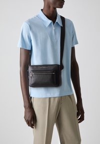 Schwarze, strukturierte Leder-Crossbody-Tasche mit zwei Reißverschlussfächern, getragen über einem hellblauen Poloshirt, kombiniert mit beigen Hosen.