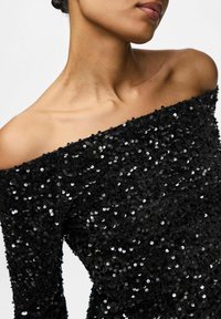 Femme portant une robe à sequins noire, décolletée, mettant en valeur ses clavicules et une partie de son visage, sur un fond uni.