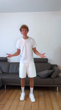 Jaunas žmogus vilkintis baltą marškinėlį, baltus šortus ir baltas sportines batus, stovi ant medinių grindų šalia tamsiai pilkos sofkos su pagalvėmis.