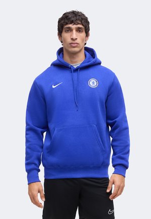 Tânăr purtând un hanorac albastru Chelsea FC cu logo Nike, stând în fața unui fundal simplu și luminos.