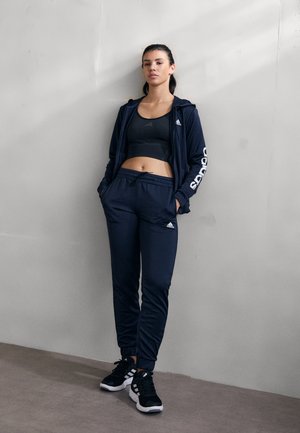 Conjunto deportivo navy que consta de una sudadera con capucha y pantalones deportivos, con logos de Adidas, combinado con zapatillas deportivas negras con detalles en blanco.
