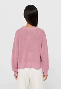Femme aux longs cheveux foncés portant des boucles d'oreilles en or, un pull rose en tricot ample et un pantalon blanc, vue de dos sur un fond uni.