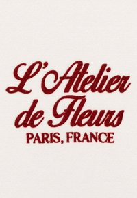 Testo ricamato su sfondo bianco: “L'Atelier de Fleurs Paris, Francia” in font corsivo bordeaux, elegante e con trama morbida.
