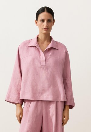 Žena oblečená v voľnom ružovom ľanovom tričku s dlhými rukávmi a golierom, s širokými rukávmi, a zladených ružových ľanových nohaviciach stojaca pred jednoduchým pozadím.