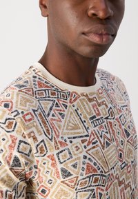 Grafisk T-shirt i en cremet farve med flerfarvede geometriske mønstre i røde, sorte og gyldne nuancer; rund hals med kontrasterende trim.
