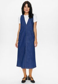 Denim overall obleka v temno modri barvi z V-izrezom, žepi na straneh in kontrastnimi šivi. V kombinaciji z belo srajco in črnimi čevlji.