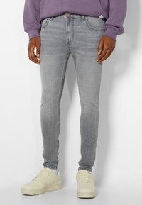 Hombre vestido con jeans ajustados de color gris desgastado, zapatillas crema y un sudadera morada, apoyado contra una pared blanca.