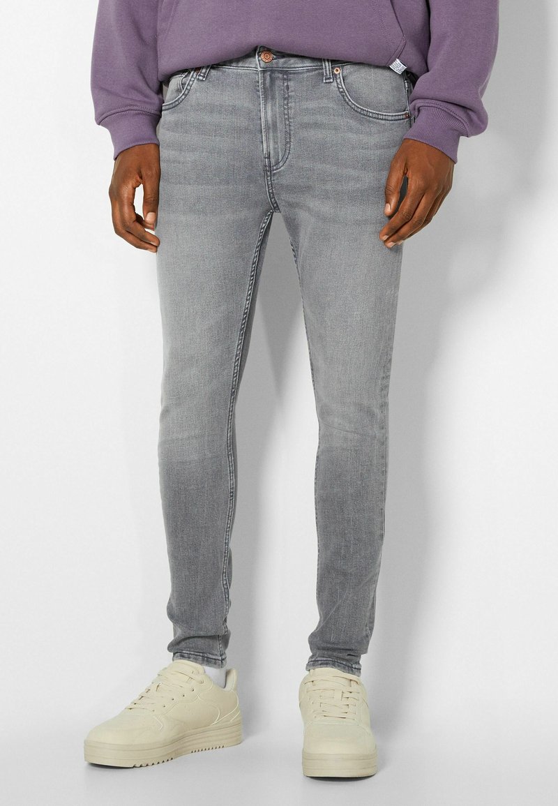 Hombre vestido con jeans ajustados de color gris desgastado, zapatillas crema y un sudadera morada, apoyado contra una pared blanca.