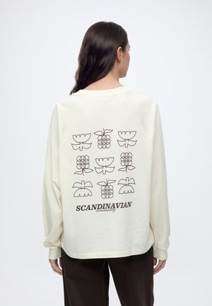 Femme aux longs cheveux foncés portant une chemise crème affichant une impression au dos de formes florales géométriques et le texte "Scandinavian community."