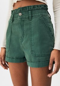 Pantalones cortos de mezclilla verde de cintura alta con dobladillos enrollados y bolsillos delanteros, combinados con un top corto de manga larga acanalado blanco.