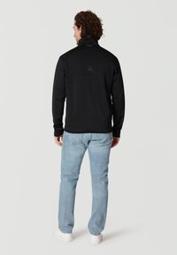 Schwarze Fleecejacke mit hohem Kragen, ausgestattet mit einem strukturierten Rückenteil und einem Logodetail, kombiniert mit hellblauen Jeans und weißen Sneakern.