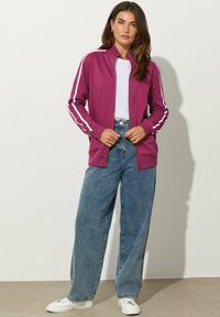 Chaqueta morada con cremallera y rayas blancas en las mangas, llevada sobre una camiseta blanca, combinada con jeans de mezclilla azul de cintura alta y zapatillas blancas.