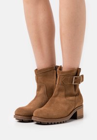 Botines de ante marrón con tacón grueso, con cremallera lateral y un detalle de hebilla ajustable en el tobillo. Superficie texturizada y suela resistente.