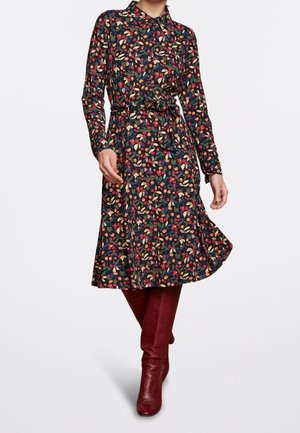 Kvinde iført en langærmet blomsterkjole med bindebånd i taljen kombineret med mørkerøde knæstøvler, der står på en ensfarvet baggrund.