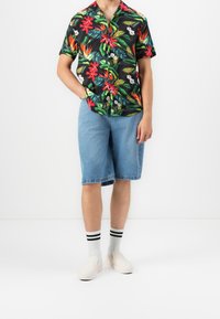 Homme portant une chemise tropicale à manches courtes noire à motifs floraux, un short en jean bleu clair, des chaussures blanches sans lacets et des chaussettes blanches à rayures noires.