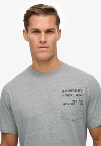 Grå crew-neck t-shirt lavet af bomuld, med en venstre brystlomme og trykt label med "SUPERDRY" tekst. Blød tekstur og afslappet pasform.