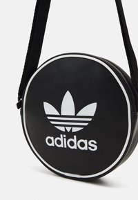 Kulatá černá kožená taška přes rameno s bílými akcenty, vyznačující se výrazným logem Adidas a designem tří vodorovných pruhů.