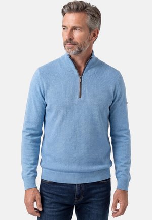 Mand med gråt hår og skæg iført en lyseblå tekstureret quarter-zip trøje og mørkeblå jeans, der kigger mod venstre.