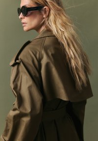 Oliveļļas zaļš tranšeju mētelis ar drapētu dizainu un jostu uz vidukļa. Modelis valkā melnas apaļas saulesbrilles, mati ieveidoti brīvās viļņos.