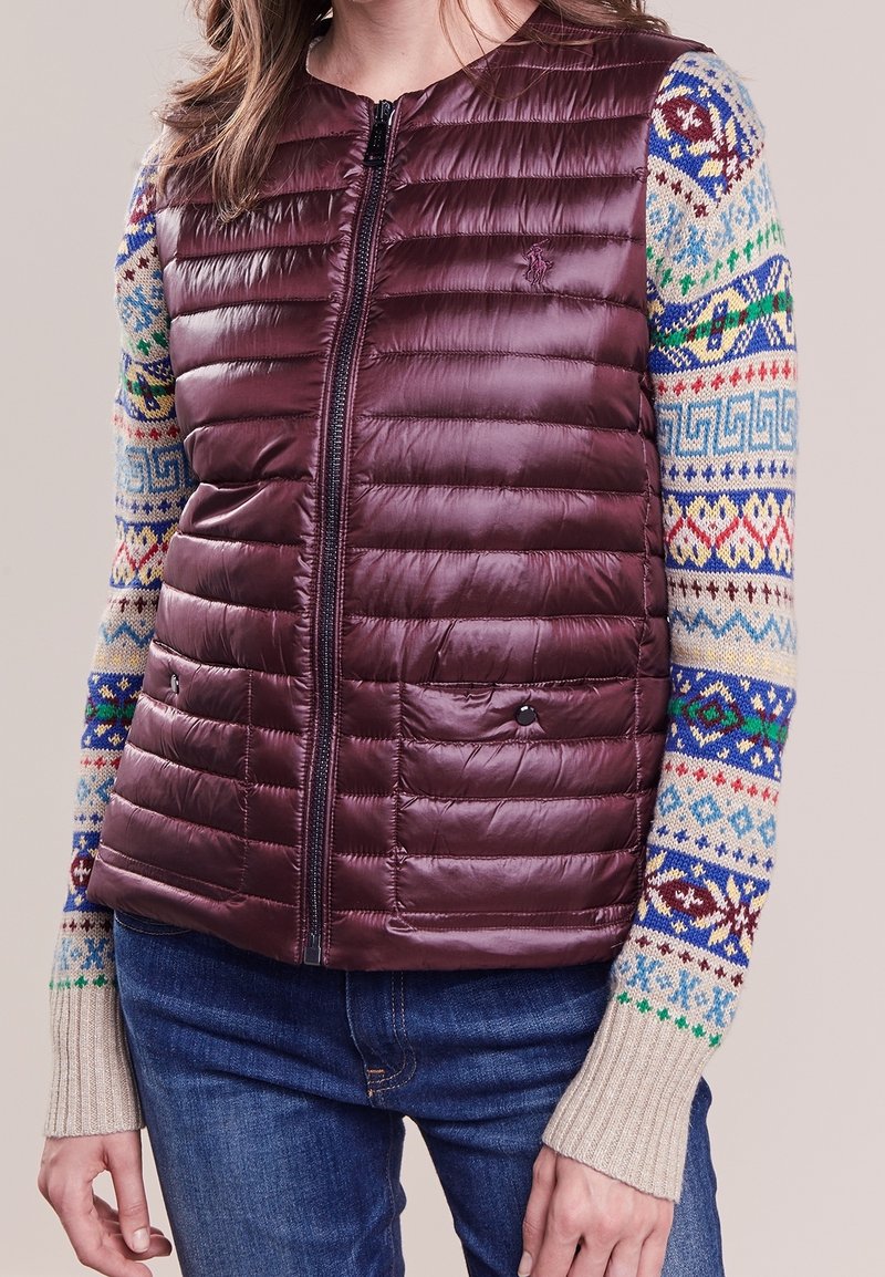 Vrouw draagt een bordeauxrode gewatteerde zip-up bodywarmer over een meerkleurige gebreide trui met patroon en blauwe spijkerbroek, staand met ontspannen armen.