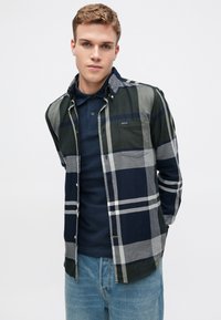 Kockás ing zöld, tengerészkék, szürke és fehér mintákkal. Gombos kialakítás, mellzsebbel, egy tengerészkék pólóra rétegezve. Világos farmernadrág.