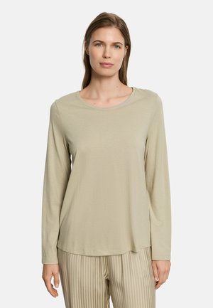 Vrouw met steil bruin haar, gekleed in een beige top met lange mouwen en beige gestreepte broek, staand tegen een effen witte achtergrond.
