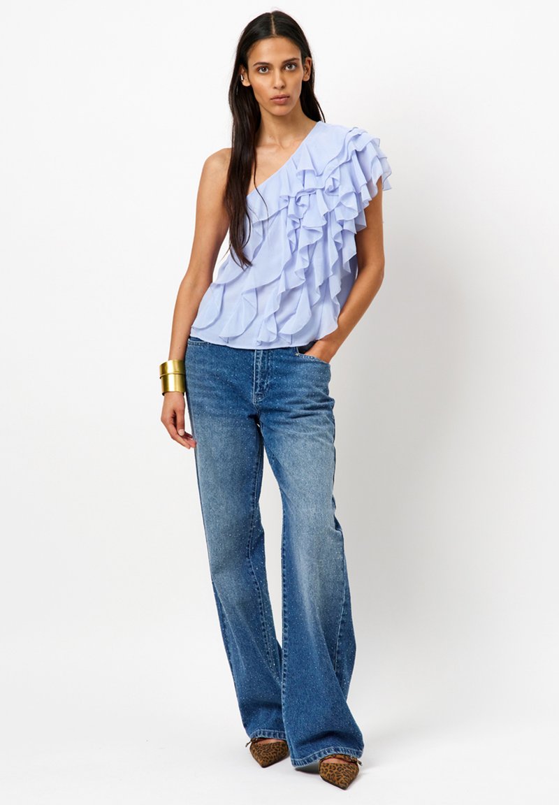 Lichtblauw ruffle-top met asymmetrisch ontwerp, gecombineerd met flare denim jeans. Textuur: gladde stof. Accessoires: gouden manchette-armband.