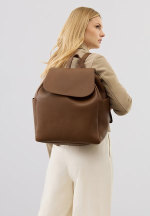 Blonde woman wearing beige jacket and cream pants carries a large brown leather backpack, facing sideways against a light background.