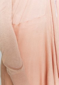 Měkký, vroubkovaný pletený cardigan v jemně broskvové barvě s dlouhými rukávy a uvolněným střihem. Obsahuje hladký povrch a jemné švy.