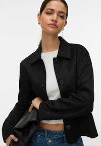 Veste courte noire en tissu suédé, avec deux gros boutons, col à revers et manches légèrement bouffantes. Associée à une pochette noire.