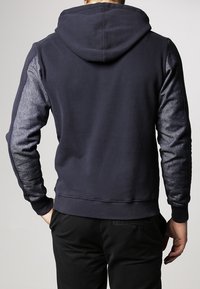 Uomo che indossa un hoodie blu navy con maniche grigie testurizzate e pantaloni neri, in piedi e rivolto all'indietro con una mano nella tasca.