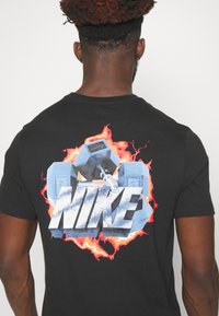 Muž v černém tričku s modrými činkami a nápisem "NIKE" v bílé a modré barvě na pozadí červeného a oranžového plamenného designu.