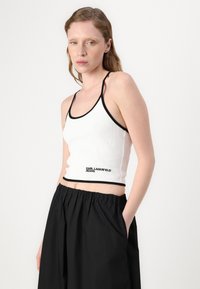 Hvit tettsittende tanktopp med svart kant og logo-tekst. Bært sammen med løstsittende svarte shorts, som viser et avslappet design.