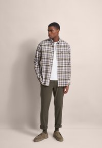 Beige, braun und weiße karierte Hemd; darunter ein weißes T-Shirt. Grüne Hose; beige Schuhe. Stehend vor einem neutralen Hintergrund.