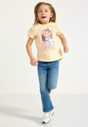 Ung pige med langt hår iført en gul "Gabby & Friends"-trøje, blå jeans og hvide sneakers, gående fremad mod en ensfarvet baggrund.