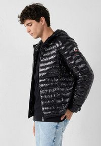 Giacca a vento nera con finitura lucida, chiusura con zip e cappuccio, caratterizzata da un design trapuntato e una patch logo sul braccio sinistro.