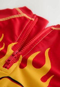 Costumul de baie roșu cu modele galbene de flăcări are un fermoar frontal în aceeași nuanță de roșu. Cusăturile sunt vizibile, evidențiind detaliile designului.