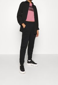 Camisa negra de botones sobre una camiseta corta rosa, combinada con pantalones de chándal negros y zapatillas negras con suelas blancas. Diseño sencillo y corte relajado.
