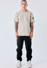 Camiseta de algodón color beige claro con un diseño gráfico circular. Combinada con pantalones cargo negros con bolsillos con cremallera y zapatillas blancas.