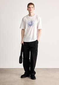 Camiseta blanca de manga corta con gráfico de sonrisa azul, combinada con pantalones cargo negros. Modelo sostiene un bolso negro de hombro. Zapatos planos.