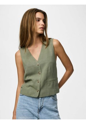 Femme portant un gilet sans manches vert olive à boutons et un jean bleu clair taille haute, debout devant un fond blanc uni.