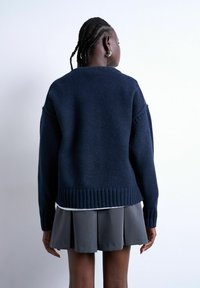 Personne aux cheveux tressés portant des boucles d'oreilles argentées en forme d'anneau, un pull bleu marine et une jupe plissée grise, debout de dos sur fond blanc.