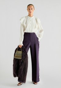 Witte gerimpelde blouse gecombineerd met diep paarse high-waisted broek. Model draagt een beaded tas en heeft een donkere bonten sjaal om. Open teen zwarte hakken.
