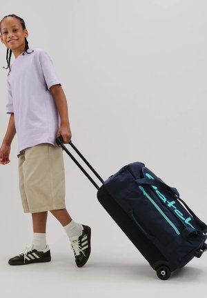 Child wearing purple shirt and beige shorts pulling dark blue wheeled duffel bag with turquoise zipper and logo.