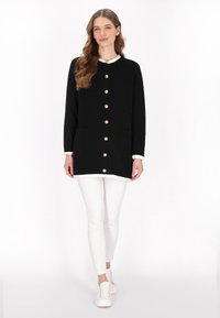 Cardigan negru, cu mâneci lungi, cu nasturi aurii, buzunare laterale și accente albe la guler și la tiv, purtat peste pantaloni și încălțăminte albă.